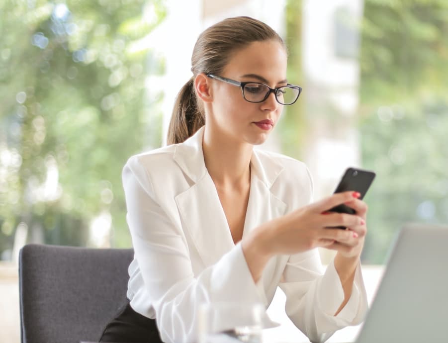 A girl in glasses sits at a table with a laptop and looks at her cell phone